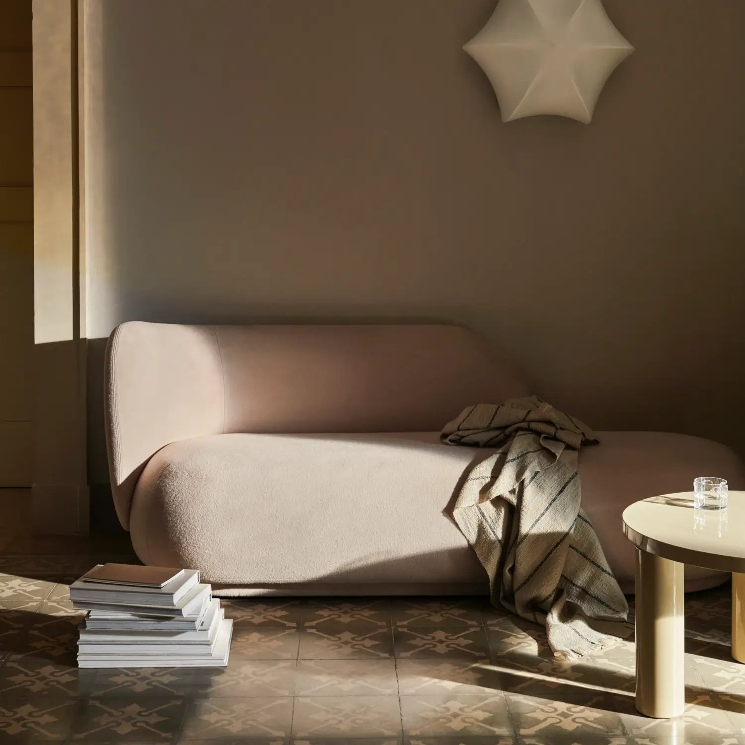 Minimalist living room featuring a softly curved, light beige sofa placed against a neutral-toned wall. A cozy, striped blanket is casually draped over one side of the sofa. Nearby, a stack of books sits on the patterned tile floor, while a sleek, round side table with a glass of water completes the serene and inviting setting. A unique, star-shaped wall fixture adds a touch of artistic flair to the calming ambiance.