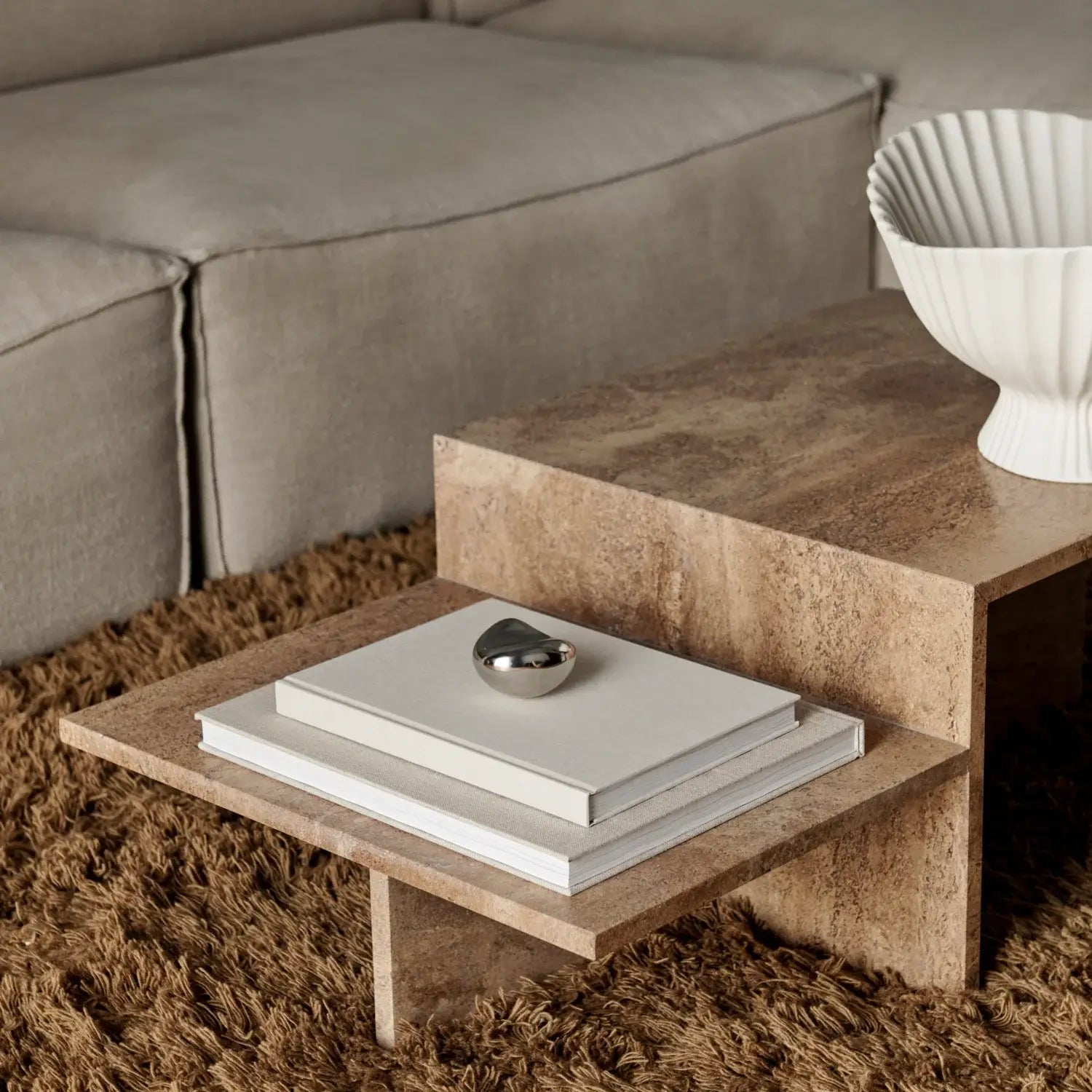 Close-up view of a modern, minimalist coffee table with a natural stone finish, featuring a stacked design. The table sits on a plush, textured brown rug, complementing the muted tones of the surrounding space. On the table’s lower tier, a neat stack of neutral-colored books is topped with a sleek, metallic decorative object. The upper tier displays a white, fluted ceramic bowl, adding a touch of elegance to the cozy yet sophisticated living area.